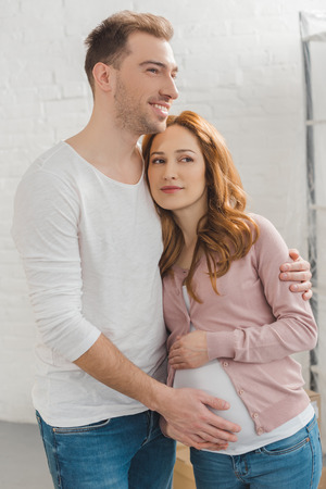 happy pregnant couple embracing and looking away at homeの写真素材