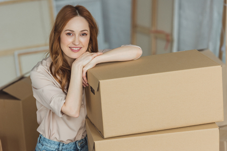 attractive smiling woman leaning on cardboard boxes at new apartment, relocation concept
