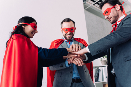 happy super businesspeople in masks and capes putting hands together in officeの写真素材