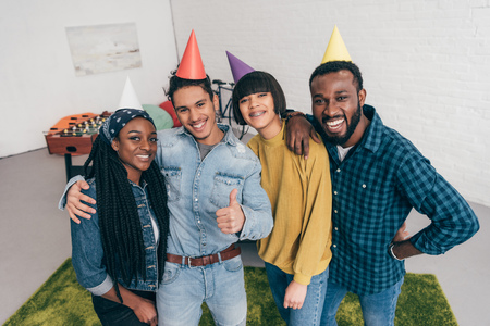 mixed race young man thumb up standing with friends in party hatsの写真素材