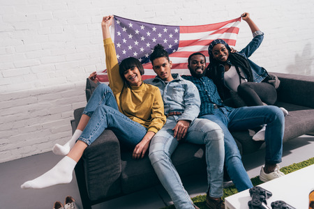 group of young multicultural friends sitting on couch with flag of USA
