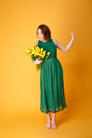 beautiful young woman in green dress looking at bouquet of yellow tulips isolated on orangeの写真素材