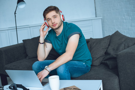 handsome young man in headphones looking at camera while working with laptop at homeの写真素材