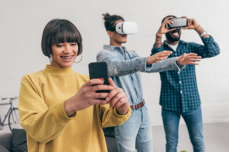 smiling mixed race young woman with smartphone and two men using virtual reality headsets behindの写真素材