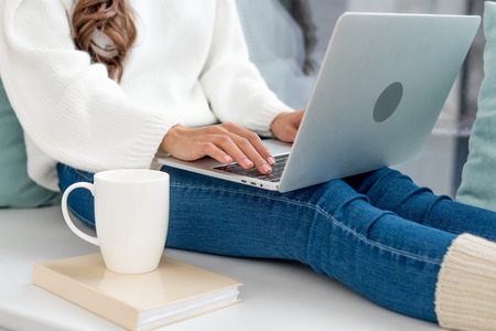 Cropped shot of freelancer with cup of coffee working with laptop on windowsill at homeの写真素材