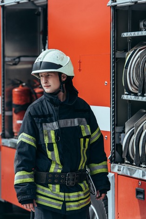 Firefighter in helmet standing at truck with water hoses inside on street