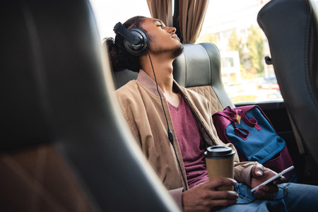 Selective focus of young mixed race man in headphones sitting with paper coffee cup and smartphone in travel busの写真素材