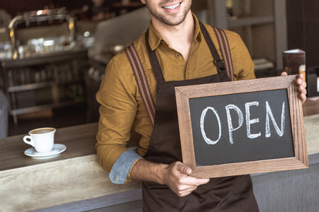 Cropped shot of happy young waiter holding chalkboard with open inscription in cafe