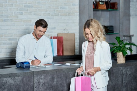 Young woman looking into shopping bags while seller taking notes in storeの写真素材