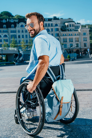 Smiling handsome man in sunglasses using wheelchair with bag on street and looking awayの写真素材