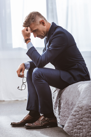 Side view of thoughtful businessman with hand on forehead sitting on bed at homeの写真素材