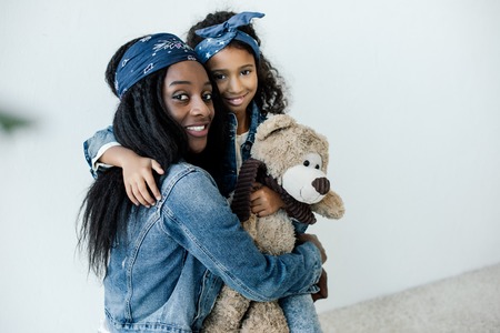 Portrait of smiling African American daughter with teddy bear hugging mother at homeの写真素材