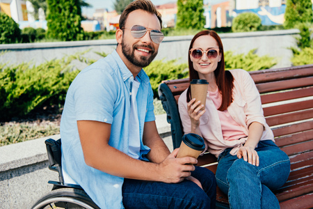 Smiling handsome boyfriend in wheelchair and girlfriend looking at camera and holding disposable coffee cups on streetの写真素材