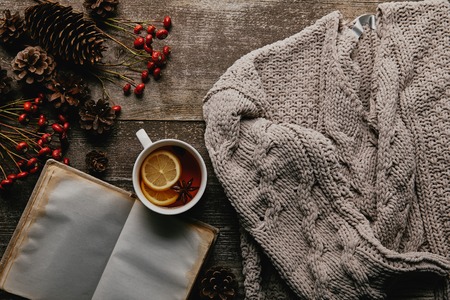 Flat lay with red holly berries, blank notebook, cup of tea and knitted sweater on wooden tabletop