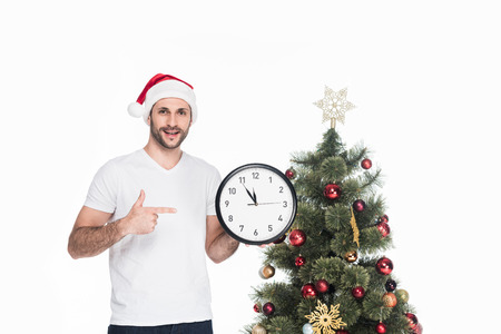 Portrait of man in Santa Claus hat pointing at clock while standing near Christmas tree isolated on...