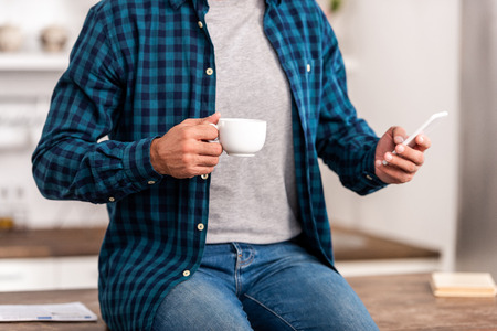 Cropped shot of man in checkered shirt holding cup of coffee and using smartphone at homeの写真素材