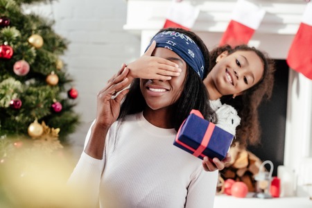 African American daughter gifting Christmas present to mother and closing her eyes for surprise at homeの写真素材