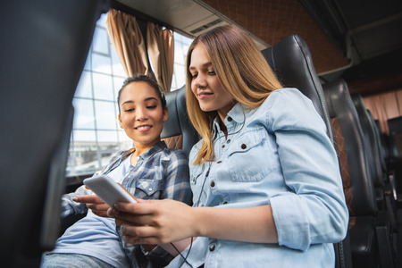 Happy young woman showing smartphone to her Asian female friend during trip on bus