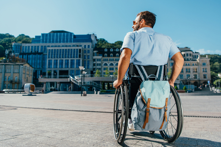Back view of man using wheelchair with bag on streetの写真素材