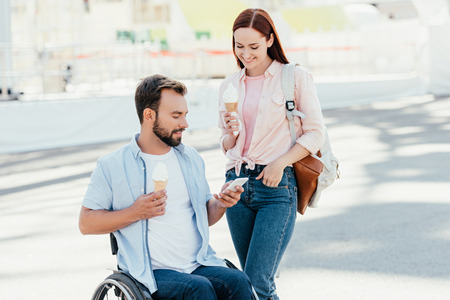 Handsome boyfriend in wheelchair and girlfriend with ice cream using smartphone on streetの写真素材