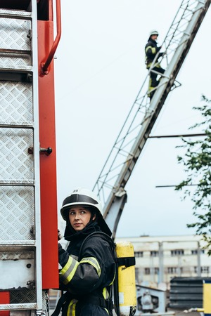 Female firefighter in uniform and helmet looking away while colleague standing on ladder on streetの写真素材