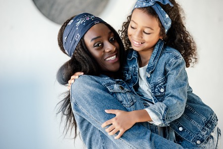 Happy African American woman holding little daughter in similar clothing on hands at homeの写真素材