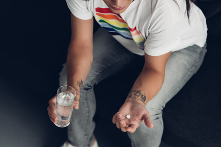 Cropped shot of young transgender man with pill and glass of water sitting on couchの写真素材
