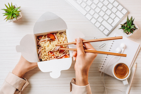 Cropped image of businesswoman eating noodle by chopsticks at table with coffee cup and computer...