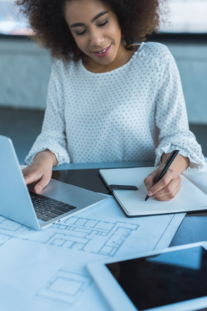 African American businesswoman using laptop and writing something to notebook in officeの写真素材