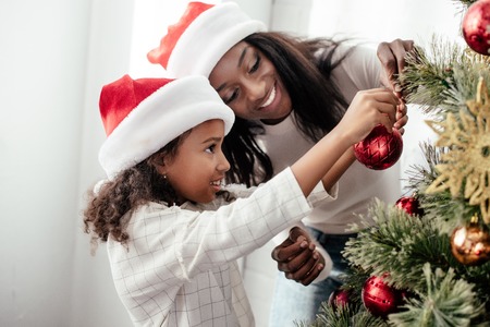 African American mother and daughter in Santa Claus hats decorating Christmas tree together at homeの写真素材