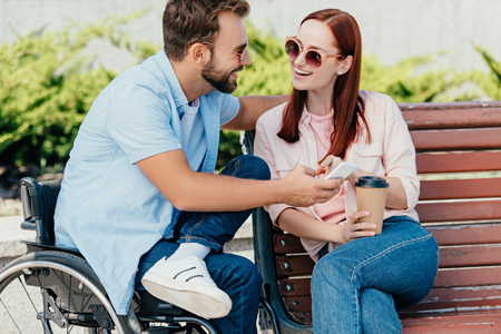 Smiling handsome boyfriend in wheelchair and girlfriend holding smartphone and looking at each other...