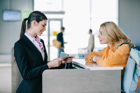 Side view of smiling airport worker checking documents of young female traveler at check-in deskの写真素材