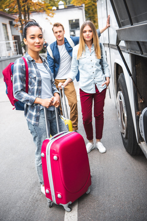 Asian young woman with wheeled bag looking at camera while her friends standing behind near travel bus at streetの写真素材