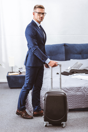 Businessman in suit and eyeglasses holding suitcase and looking at camera in bedroomの写真素材