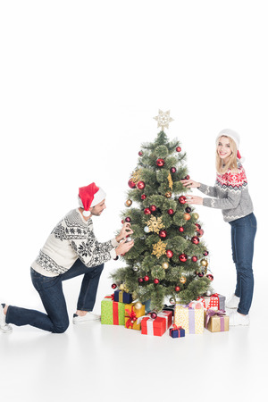 Young couple in Santa hats decorating Christmas tree together isolated on white backgroundの写真素材
