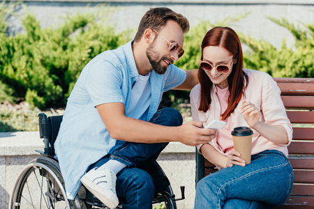 Happy handsome boyfriend in wheelchair and girlfriend looking at smartphone on street