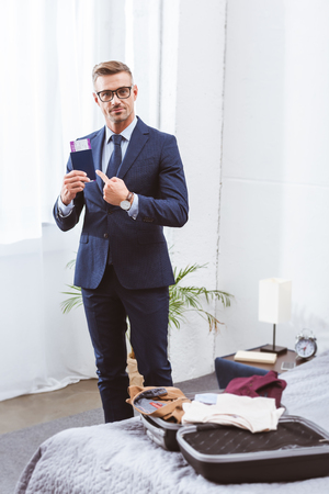 Handsome businessman holding passport with boarding pass and looking at camera while packing suitcase at homeの写真素材