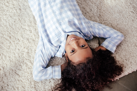 Top view of smiling adorable African American child in pajamas lying on carpet in roomの写真素材
