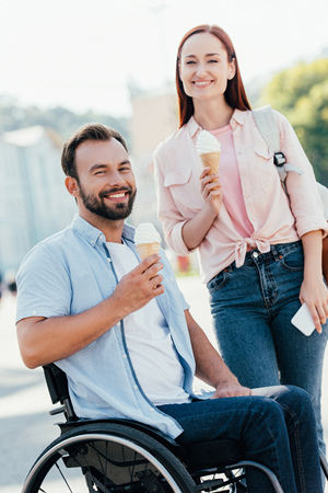 Happy handsome boyfriend in wheelchair and girlfriend with ice cream looking at camera on streetの写真素材