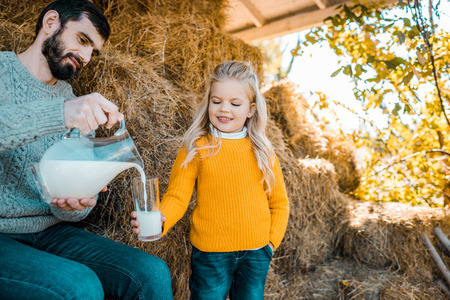 Adult female farmer pouring milk to little daughter near hay stacks at ranchの写真素材