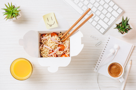 Elevated view of wok box with chopsticks, orange juice, symbol of smile and coffee cup at table in officeの写真素材