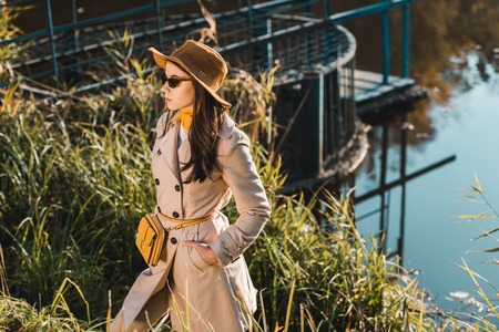 Attractive young woman  in sunglasses, trench coat and hat posing near pond in parkの写真素材