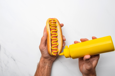 Cropped shot of man pouring mustard onto hot dog on white marble surfaceの写真素材