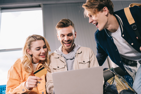 Cheerful friends with credit card using laptop in airportの写真素材