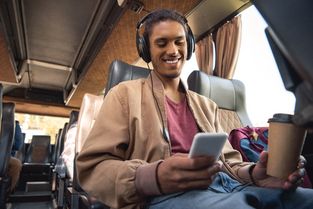 Smiling mixed race man in headphones sitting with paper coffee cup and smartphone in travel busの写真素材
