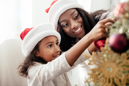 African American mother and daughter in Santa Claus hats decorating Christmas tree together at homeの写真素材