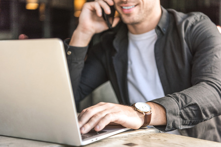 Cropped shot of smiling young freelancer working with laptop and talking by phone in cafeの写真素材