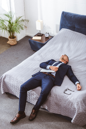 High angle view of overworked businessman holding laptop and lying on bedの写真素材