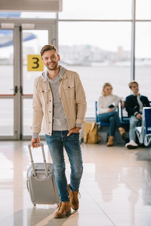 handsome young man with suitcase smiling at camera in airportの写真素材