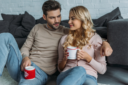 Smiling boyfriend and girlfriend holding cups of cappuccino and sitting on carpet in living roomの写真素材